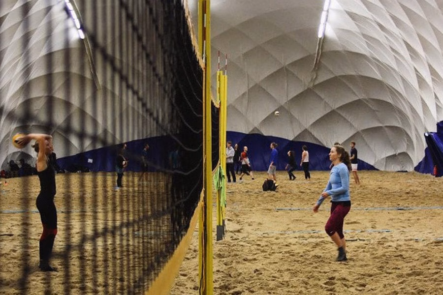 Paranet Nutzungskonzept Beachvolleyball 3 Blick durch ein Volleyballnetz auf Sandplätze und Spieler in einer hellen Traglufthalle.