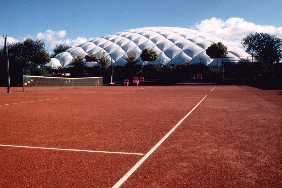 Paranet Nutzungskonzept Tennis Traglufthalle 2 Roter Tennis-Freiplatz vor der weißen Kuppel einer PARANET Traglufthalle unter blauem Himmel.