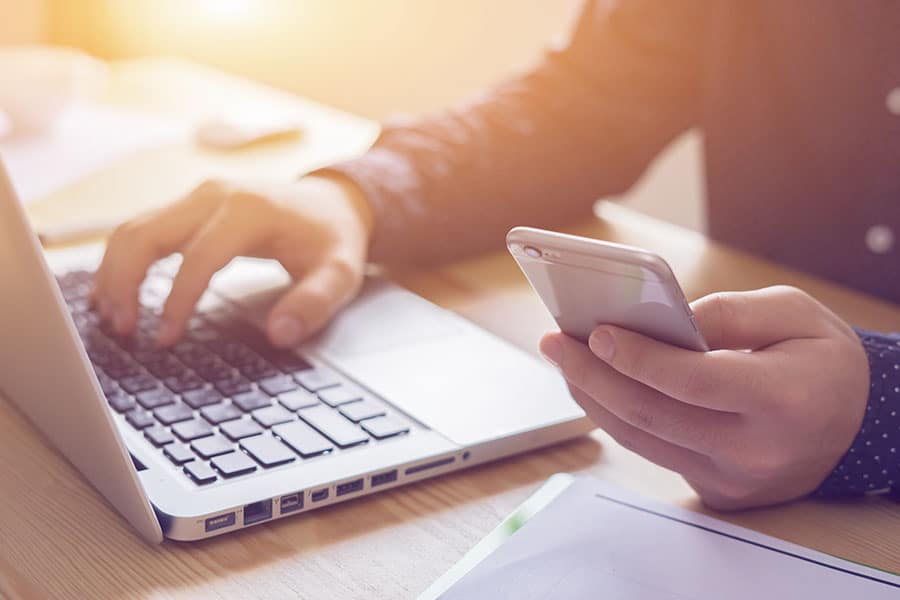 Person sitzt vor einem Laptop mit dem Handy in der Hand