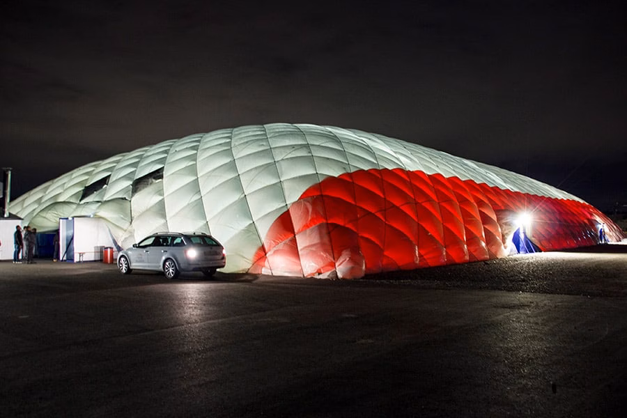Paranet Traglufthalle Galerie 8 Traglufthalle mit rot-weißer Hülle bei Nacht auf einem asphaltierten Platz mit geparktem Auto.
