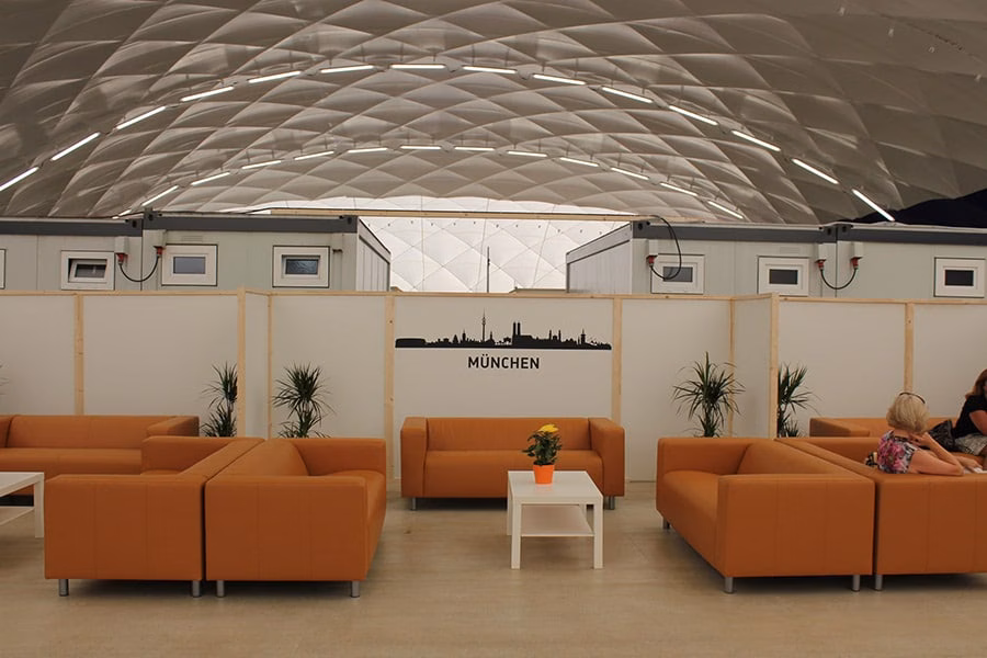 Sitzbereich mit orangen Sofas vor Trennwand mit München-Skyline im Innenraum einer Traglufthalle.