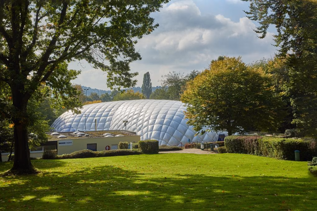 image Außenansicht einer weißen Traglufthalle, die zwischen grünen Bäumen und Wiesen in die Landschaft eingebettet ist.