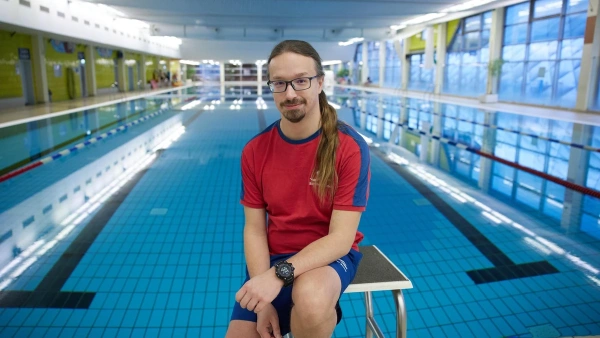 Ein Mann mit Brille und rotem Shirt sitzt auf einem Startblock am Beckenrand eines großen Hallenbads.