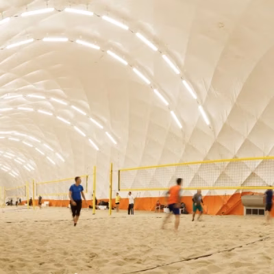 Menschen beim Beachvolleyball auf Sandplätzen in einer hell erleuchteten Traghalle.